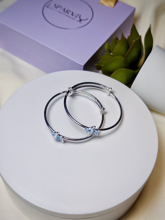 Silver baby kada with blue fish charms on a white surface with a purple box in the background.