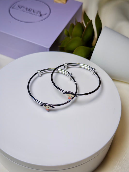 Silver baby kada with doll charms on a white surface, with a purple box and green leaves in the background.