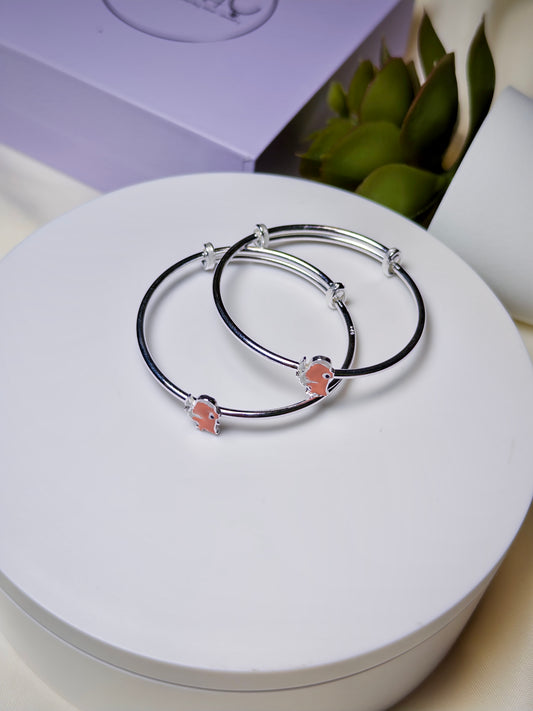 Two silver baby kada with rhino charms on a white surface with a purple box and green leaves in the background.