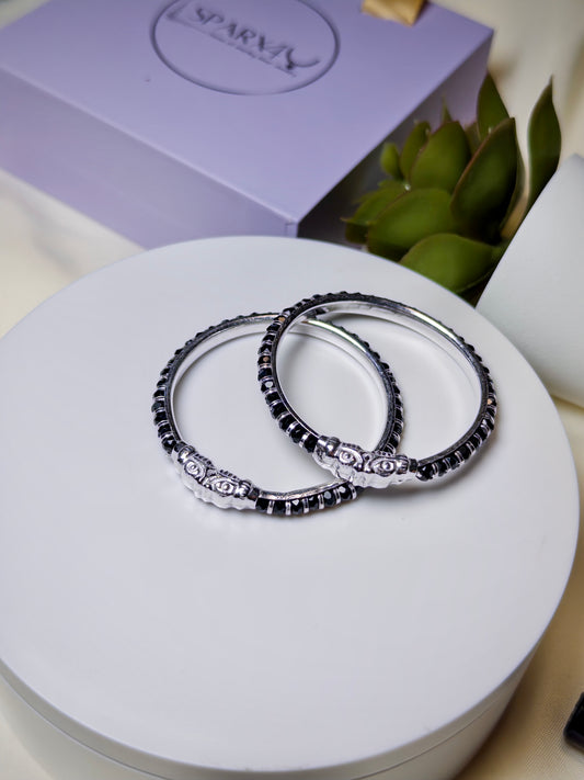 Two silver bracelets with black stones on a white surface, with a purple box and green leaves in the background.