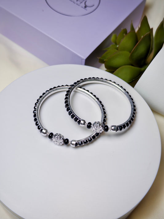 Two black beaded baby bracelets with silver accents on a white surface, with a purple box and green plant in the background.