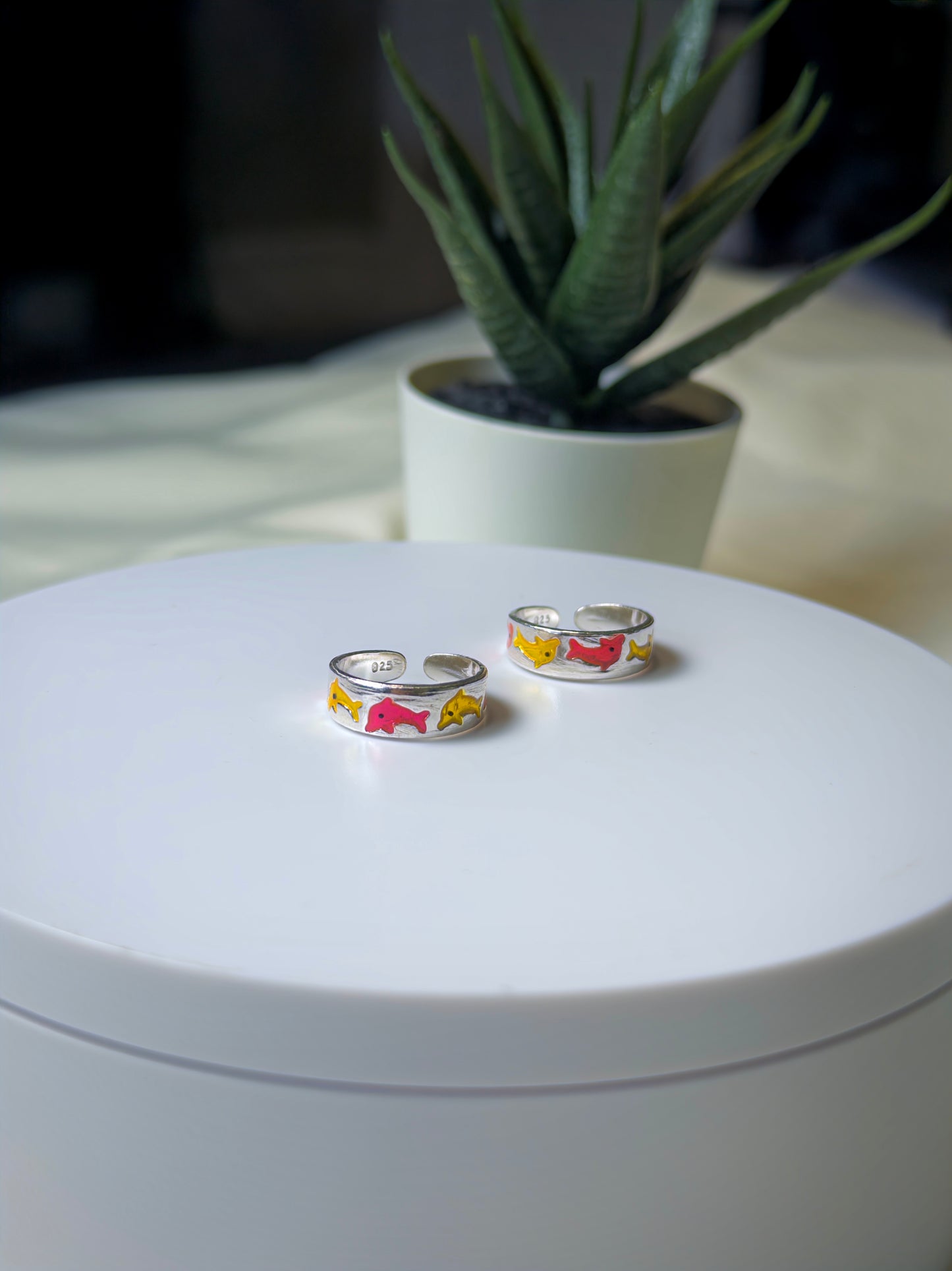Two silver toe rings with colorful designs on a white surface, with a plant in the background.