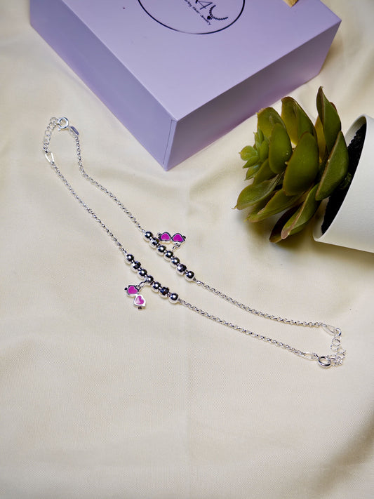 Kids silver anklet with pink glasses charm on a light surface with a plant in the background