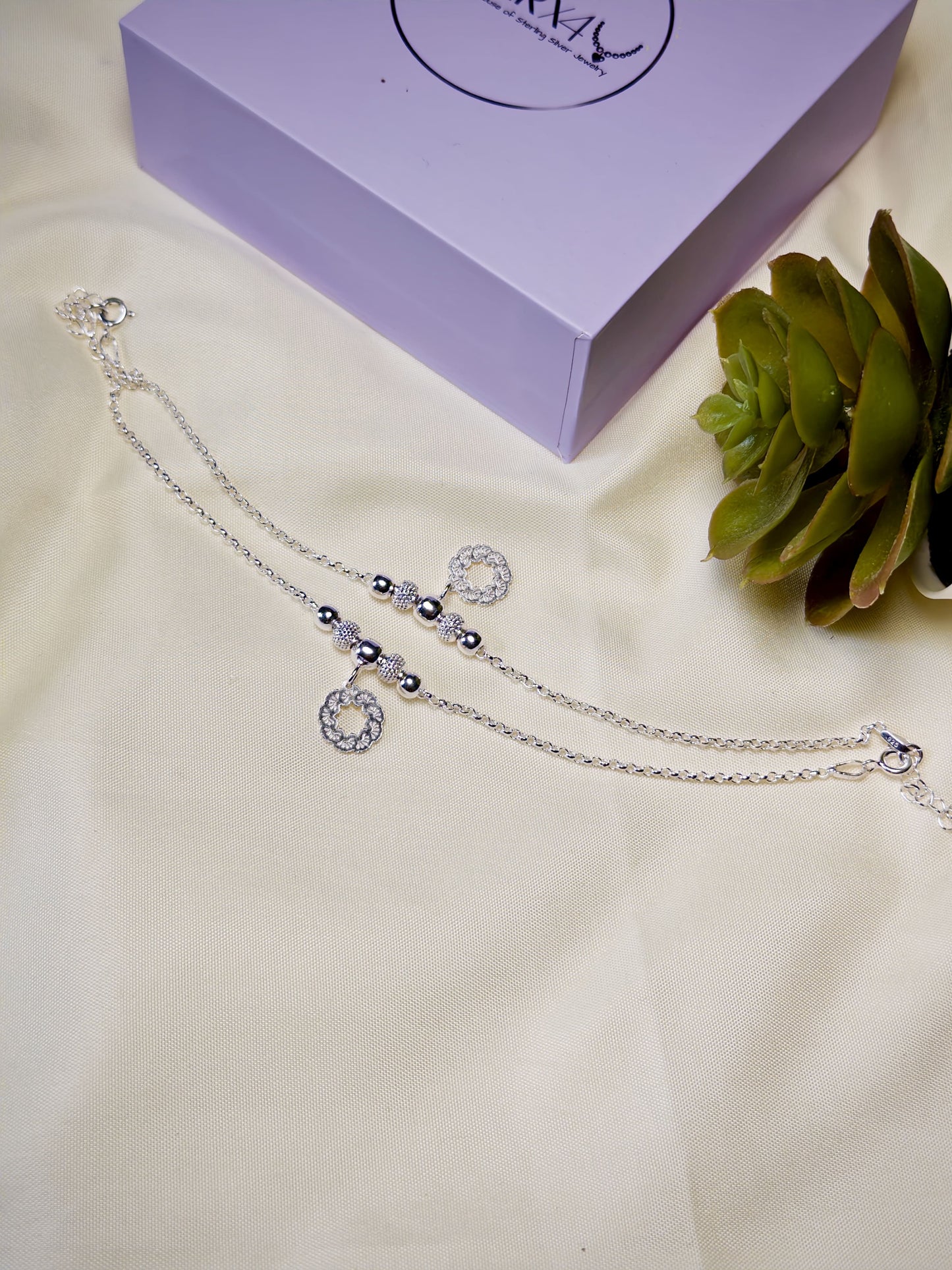 Kids silver anklets on a white surface with a purple box and green plant in the background.