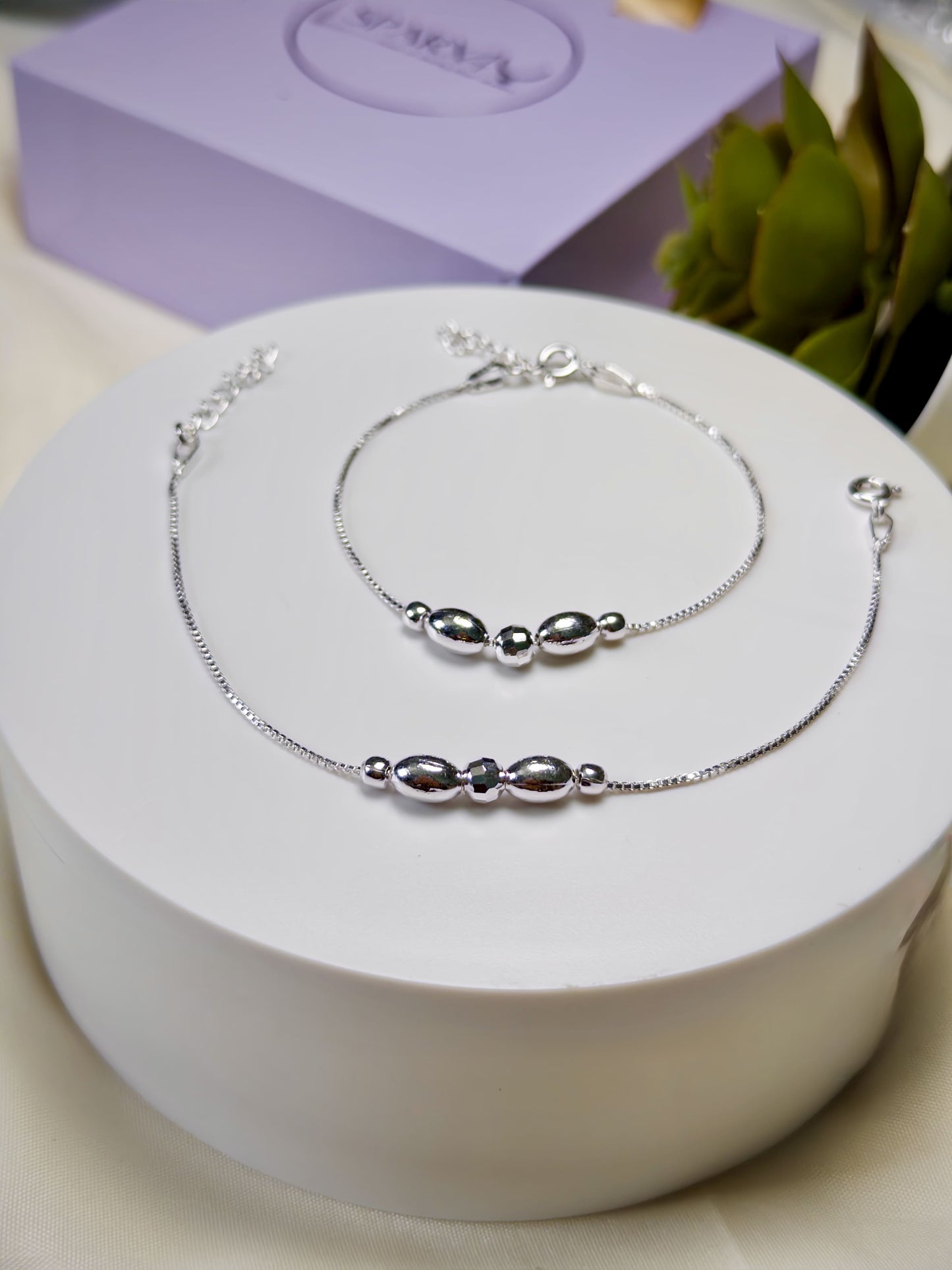 Silver anklets with silver beads on a white box with a purple box and green leaves in the background
