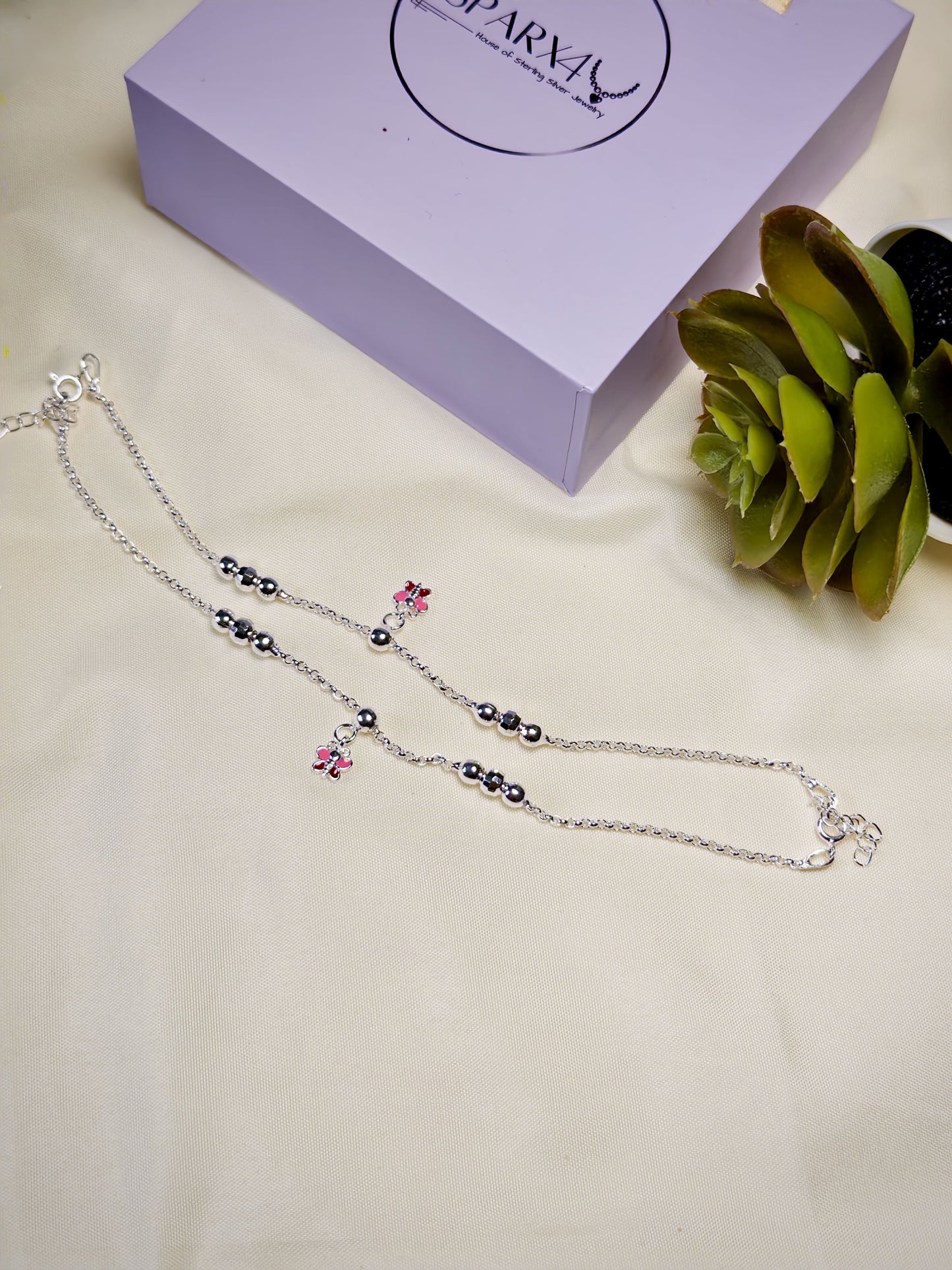 Kids silver anklets with pink butterfly on a light surface next to a purple box.