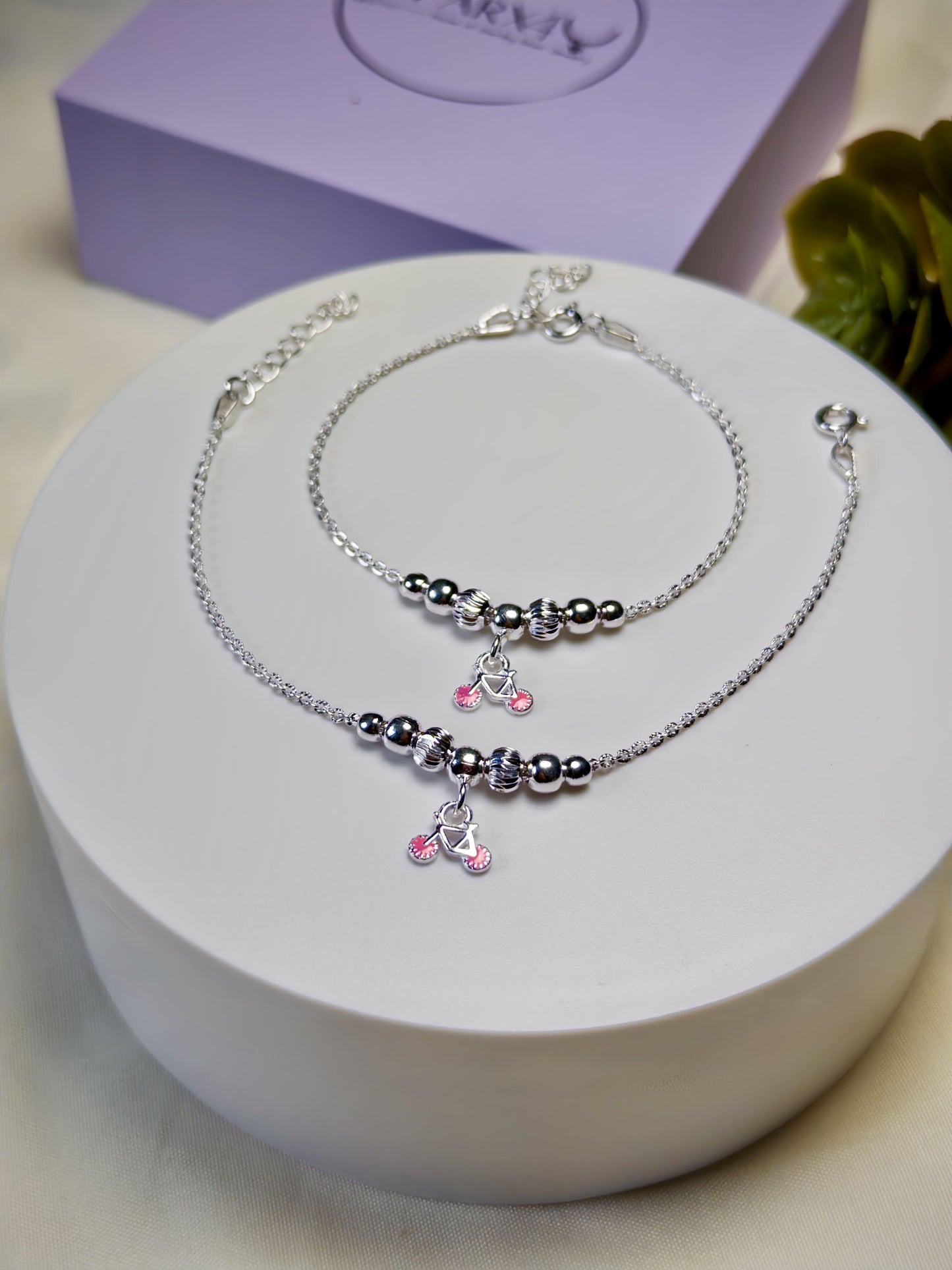 Kids silver anklets with pink cycle charms on a white surface with a purple box in the background