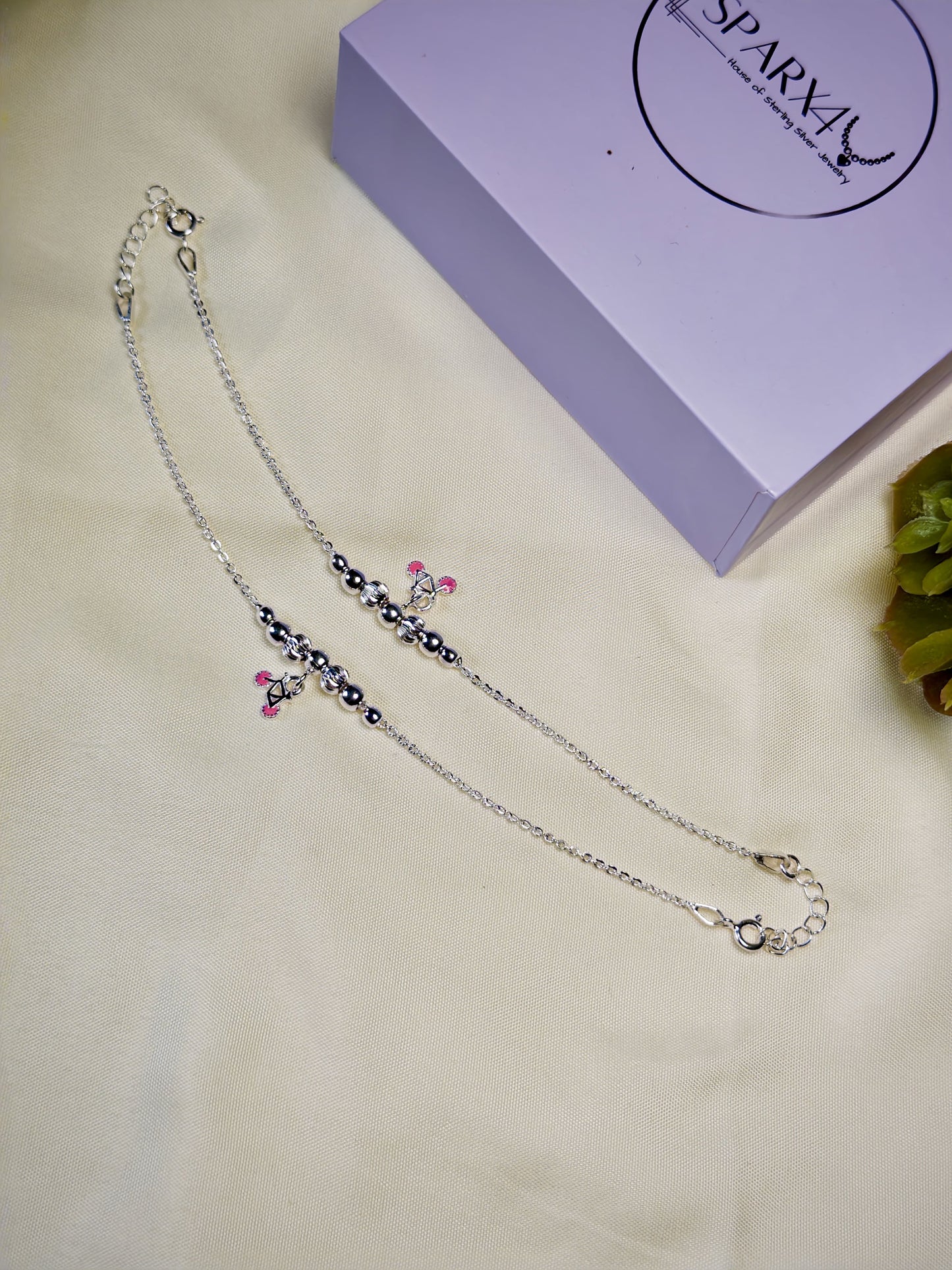 Kids silver anklets with pink charm on a beige surface with a purple box in the background