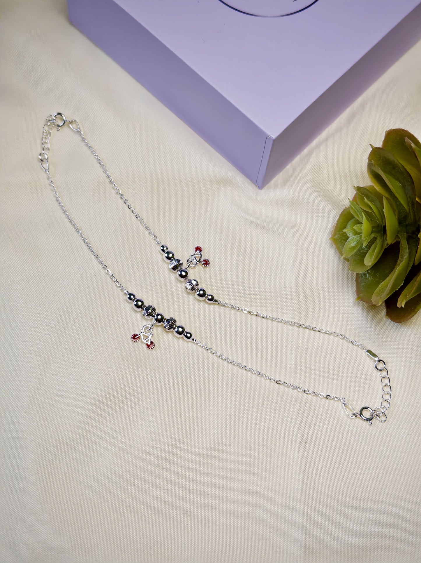 Kids silver anklets with red cycle charm on a light surface with a purple box and green plant in the background