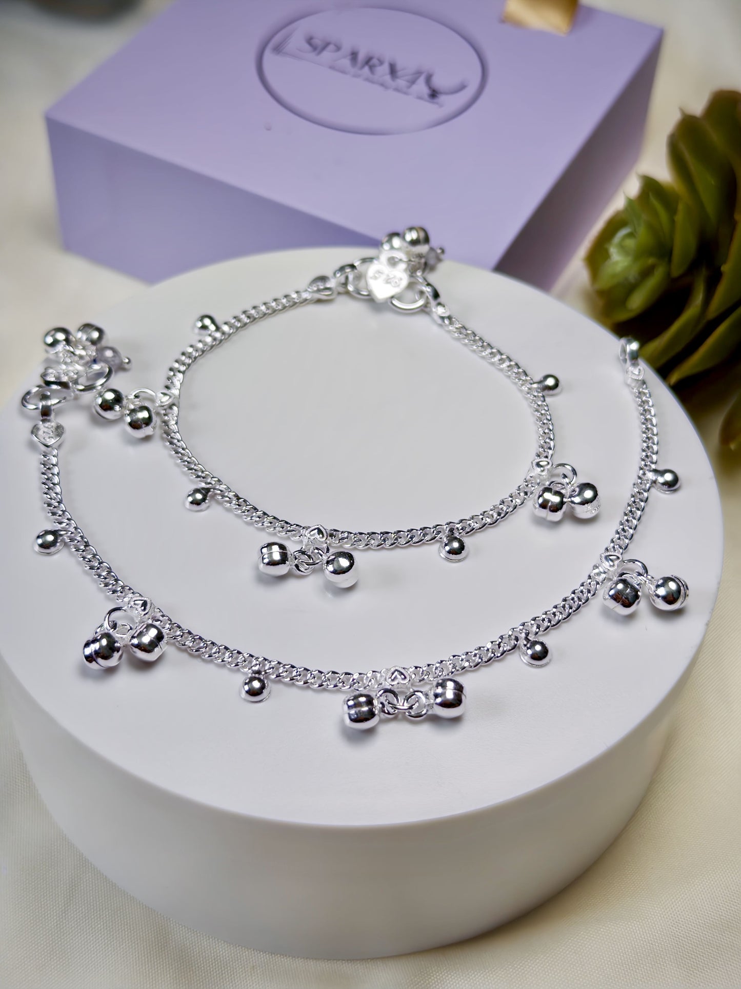 Kids silver anklets with heart-shaped charms on a white surface with a purple box in the background