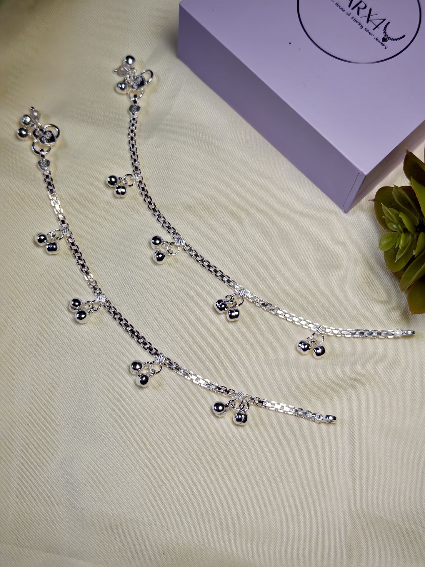 Kids silver anklets with charms on a light surface with a purple box in the background