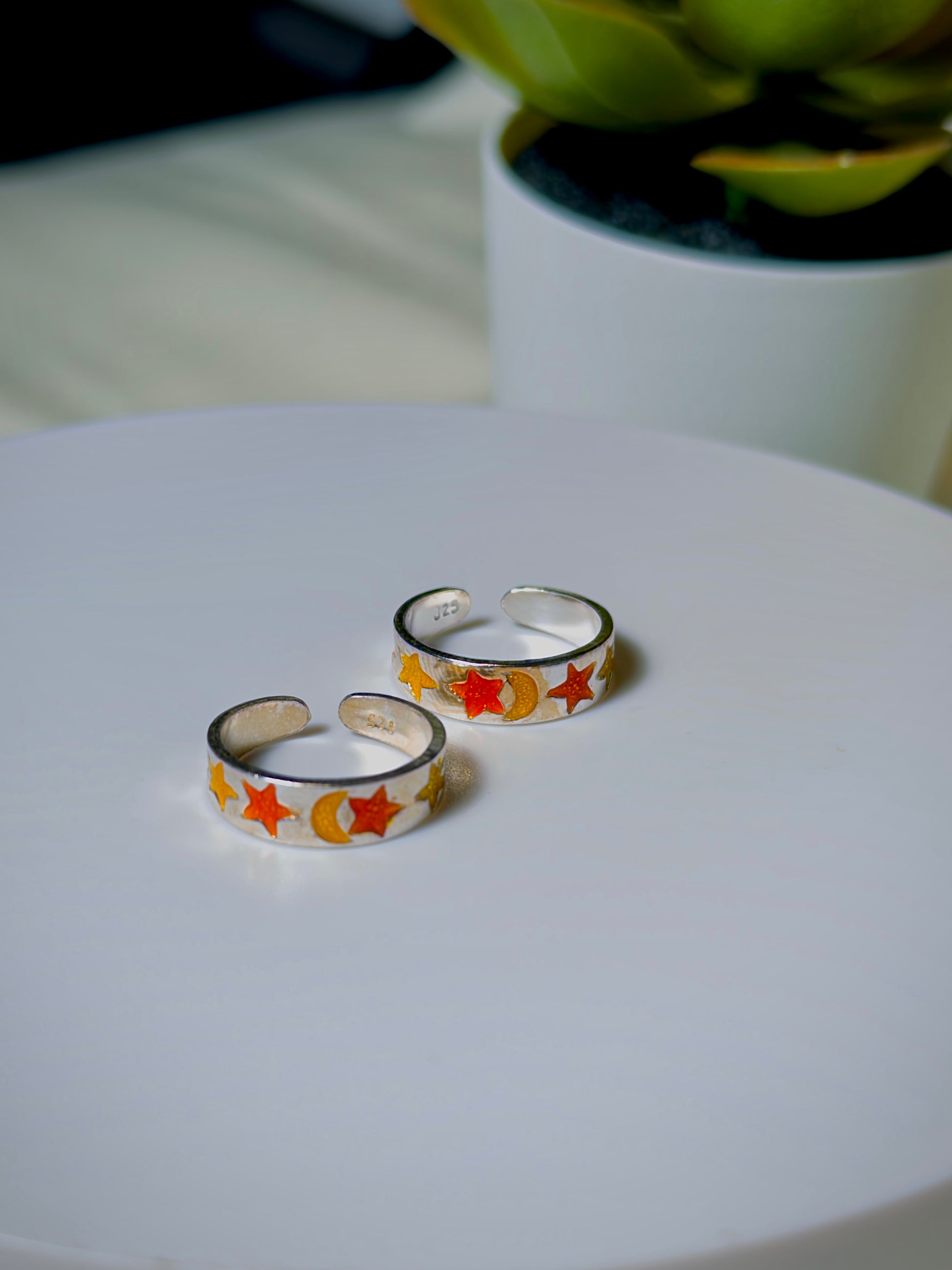 Silver toe rings with star and moon designs on a white surface, with a blurred plant in the background.
