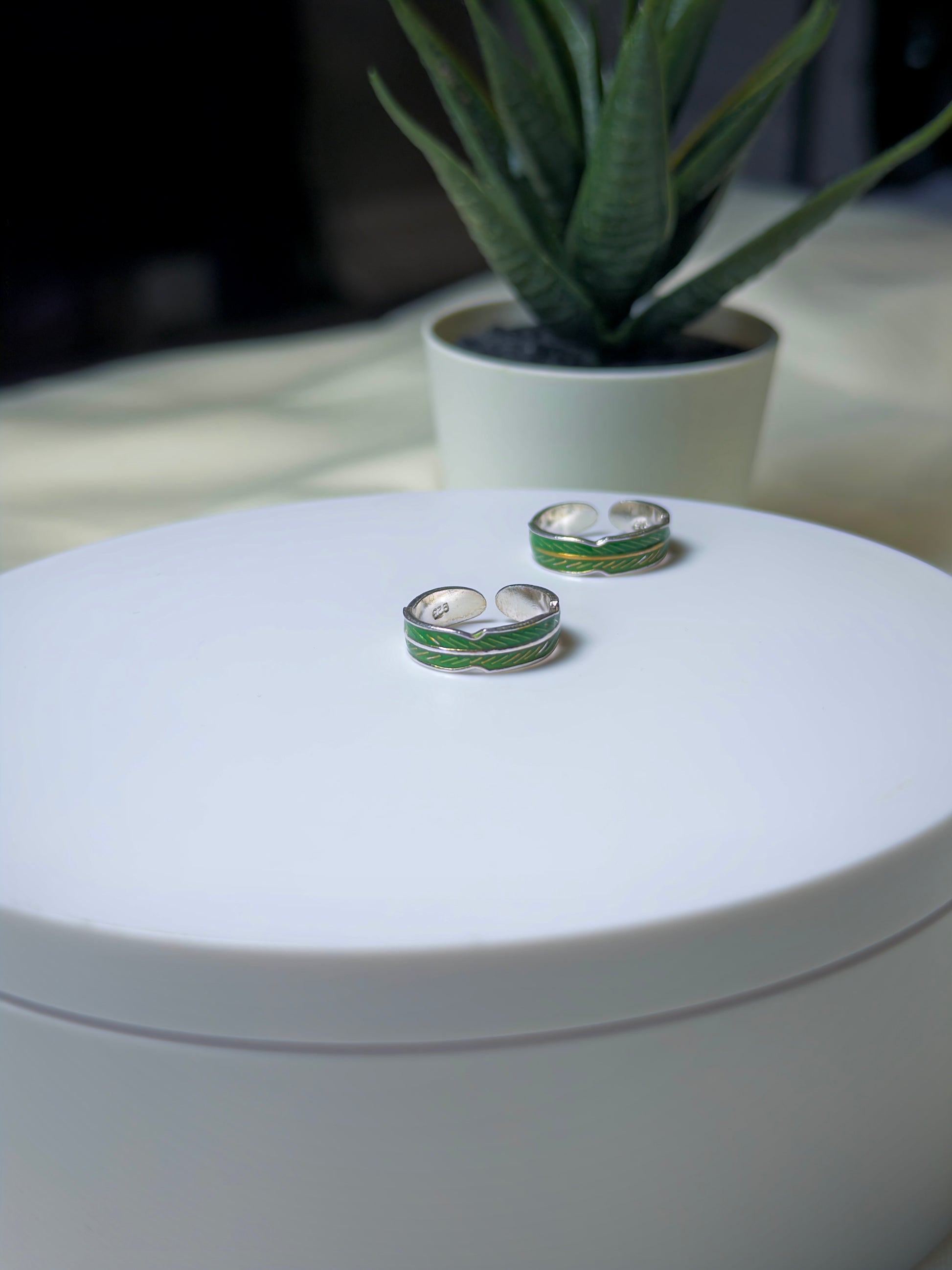 Silver toe rings with green accents on a white surface, with a plant in the background.