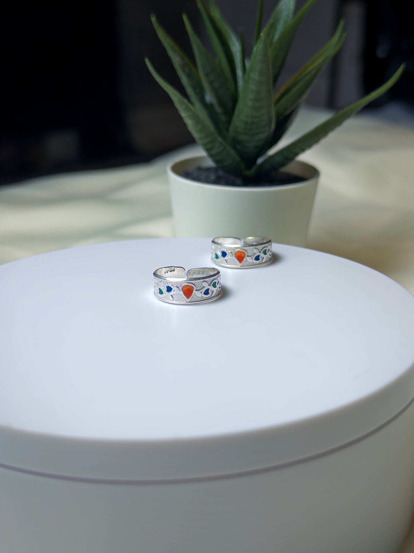 Silver toe rings with colorful stones on a white surface, potted plant in the background