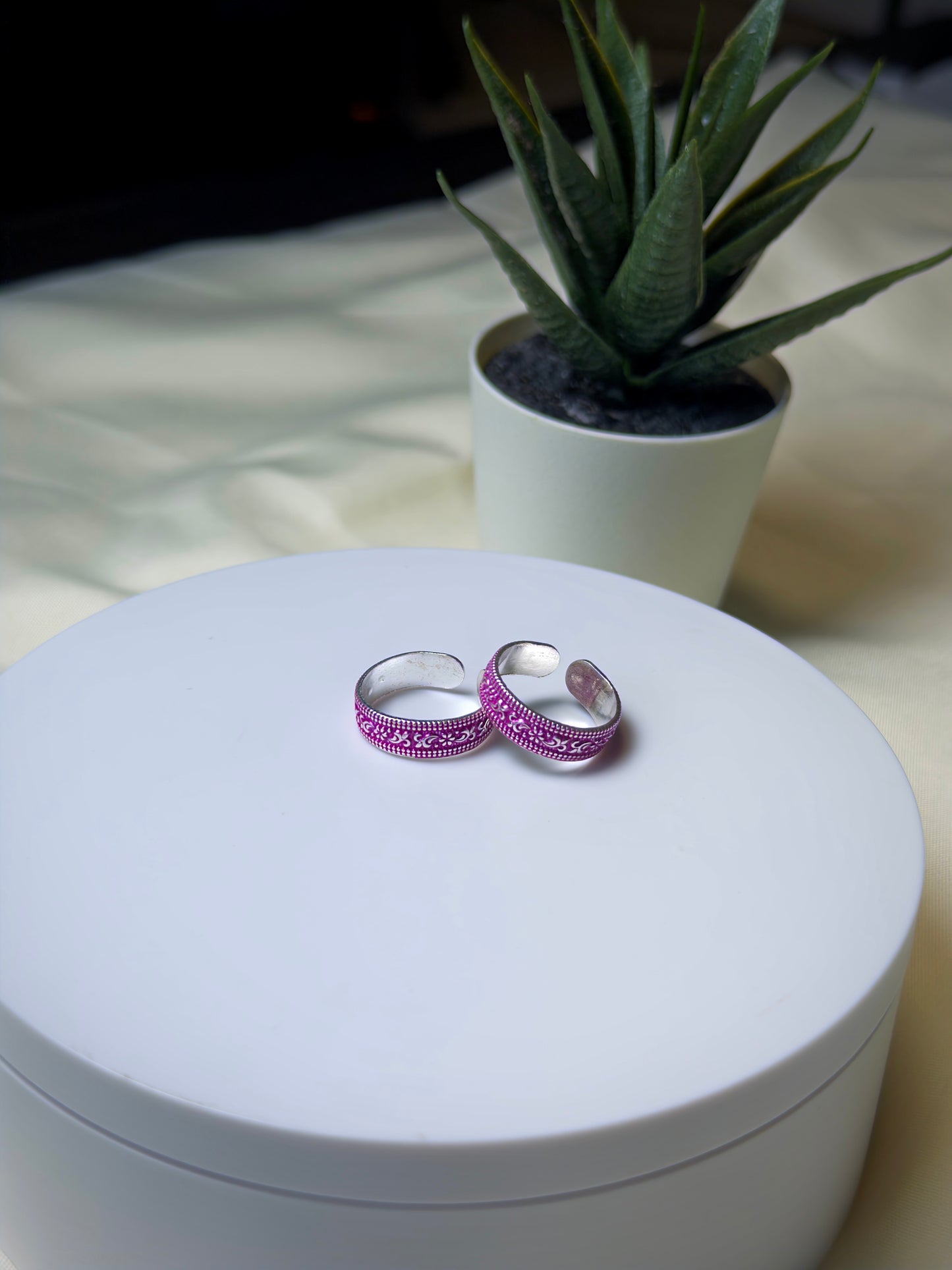 Two silver toe rings with pink gemstones on a white surface, with a plant in the background.