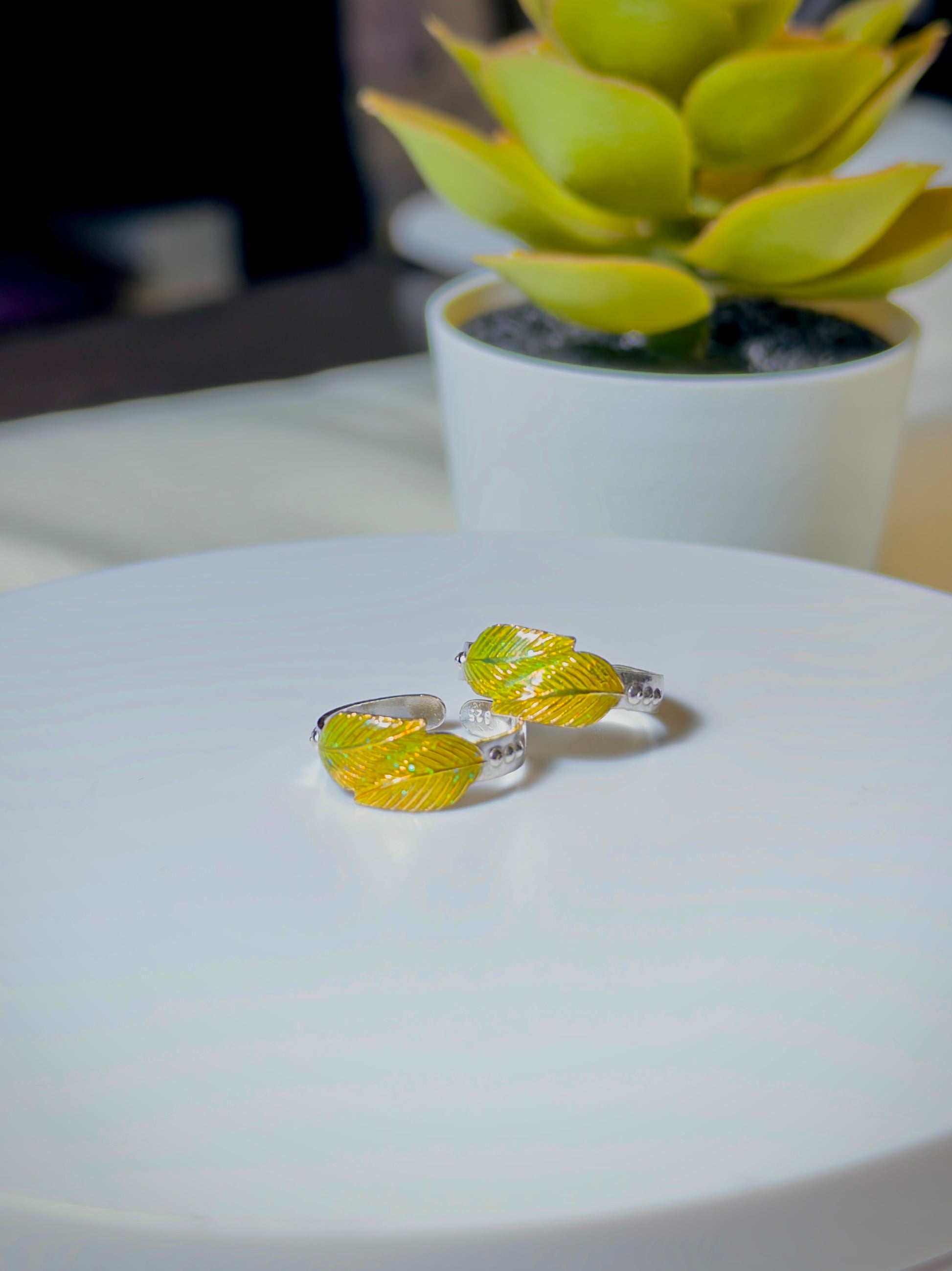 Two leaf-shaped toe rings on a white surface with a blurred plant in the background