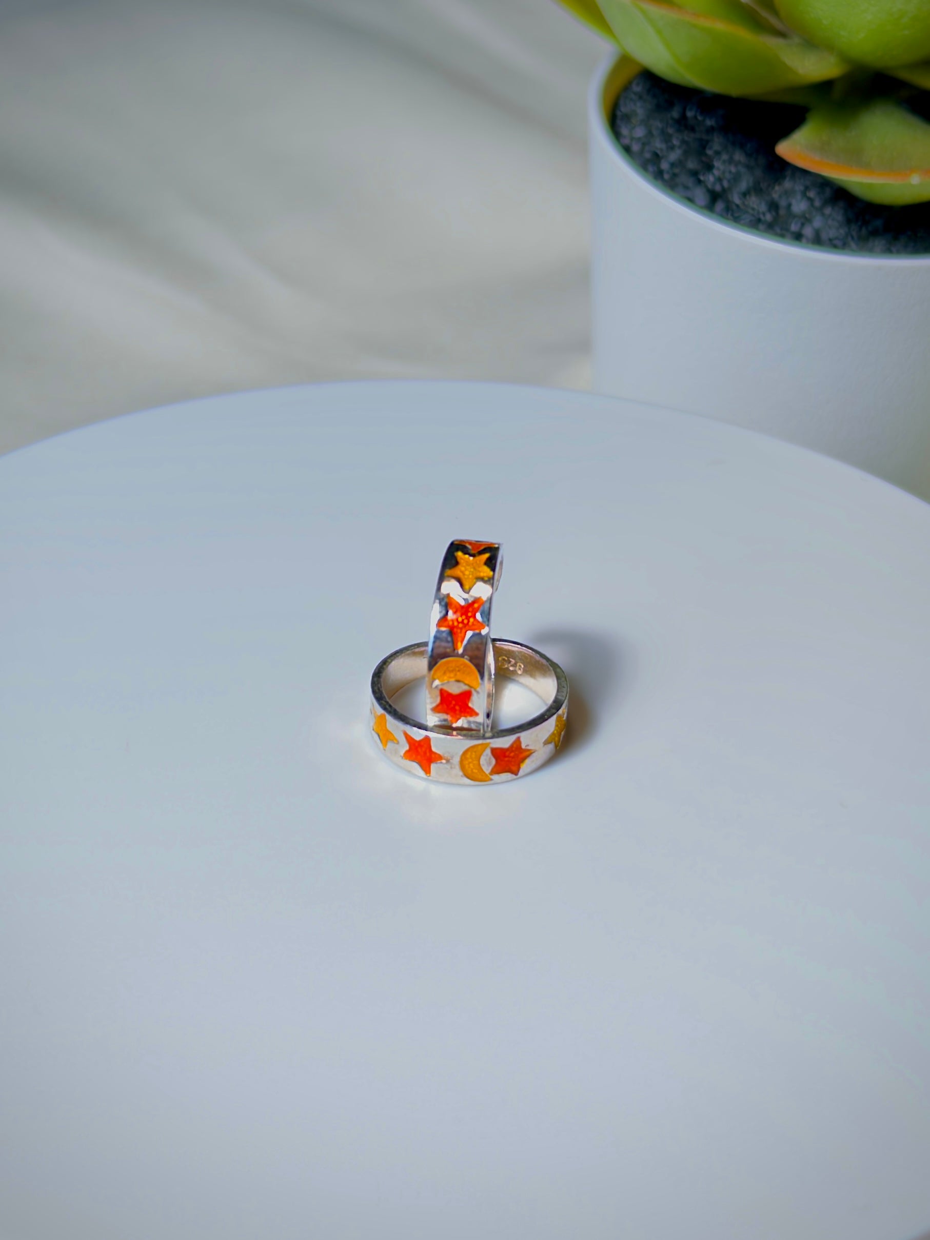 Two rings with star designs on a white surface, with a blurred plant in the background.