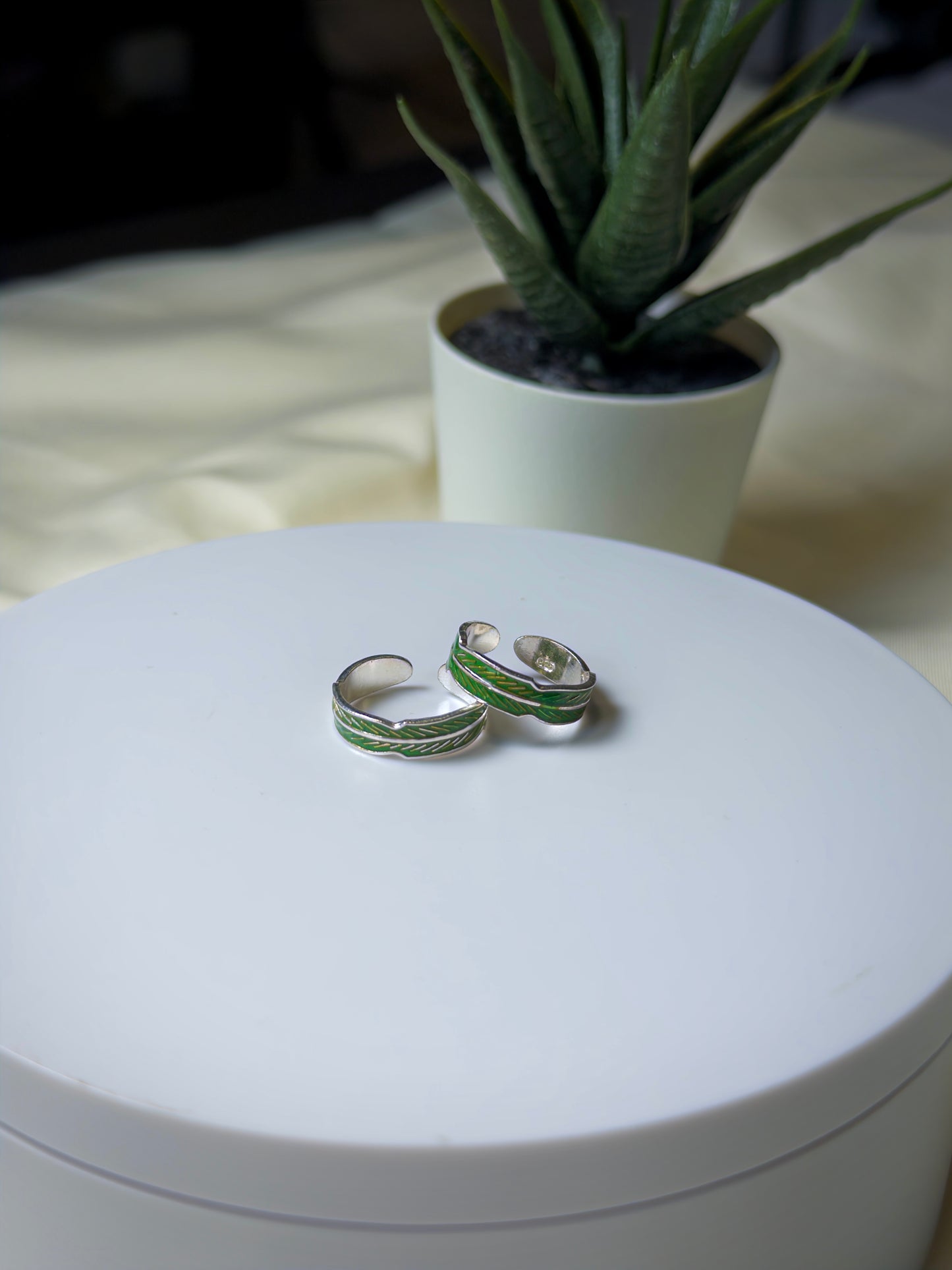 Silver toe rings with green accents on a white surface, with a potted plant in the background.