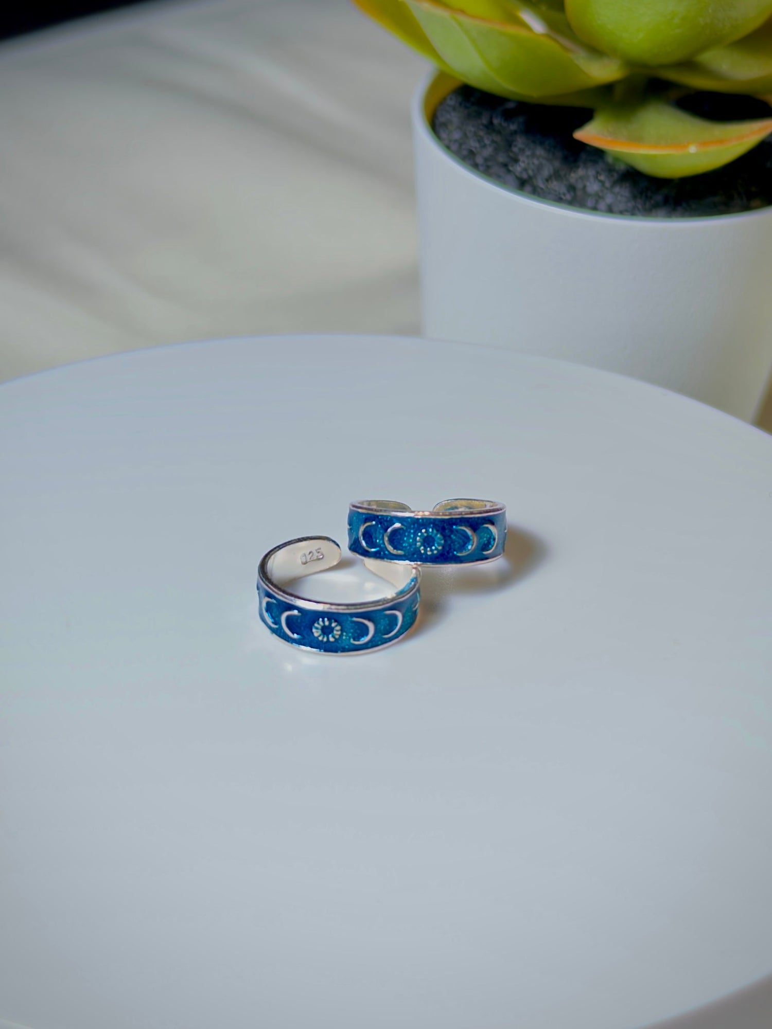 Two silver toe rings with blue engraving on a white surface, with a potted plant in the background.