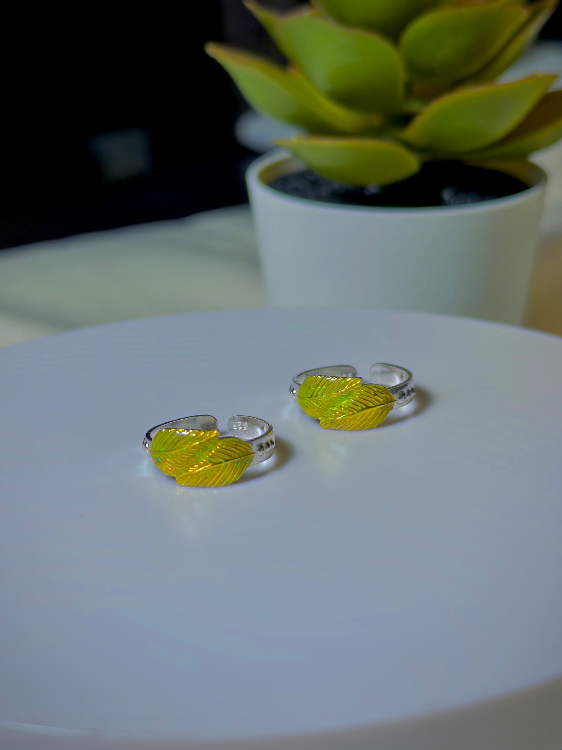 Two silver toe rings with yellow leaf designs on a white surface, with a blurred plant in the background.