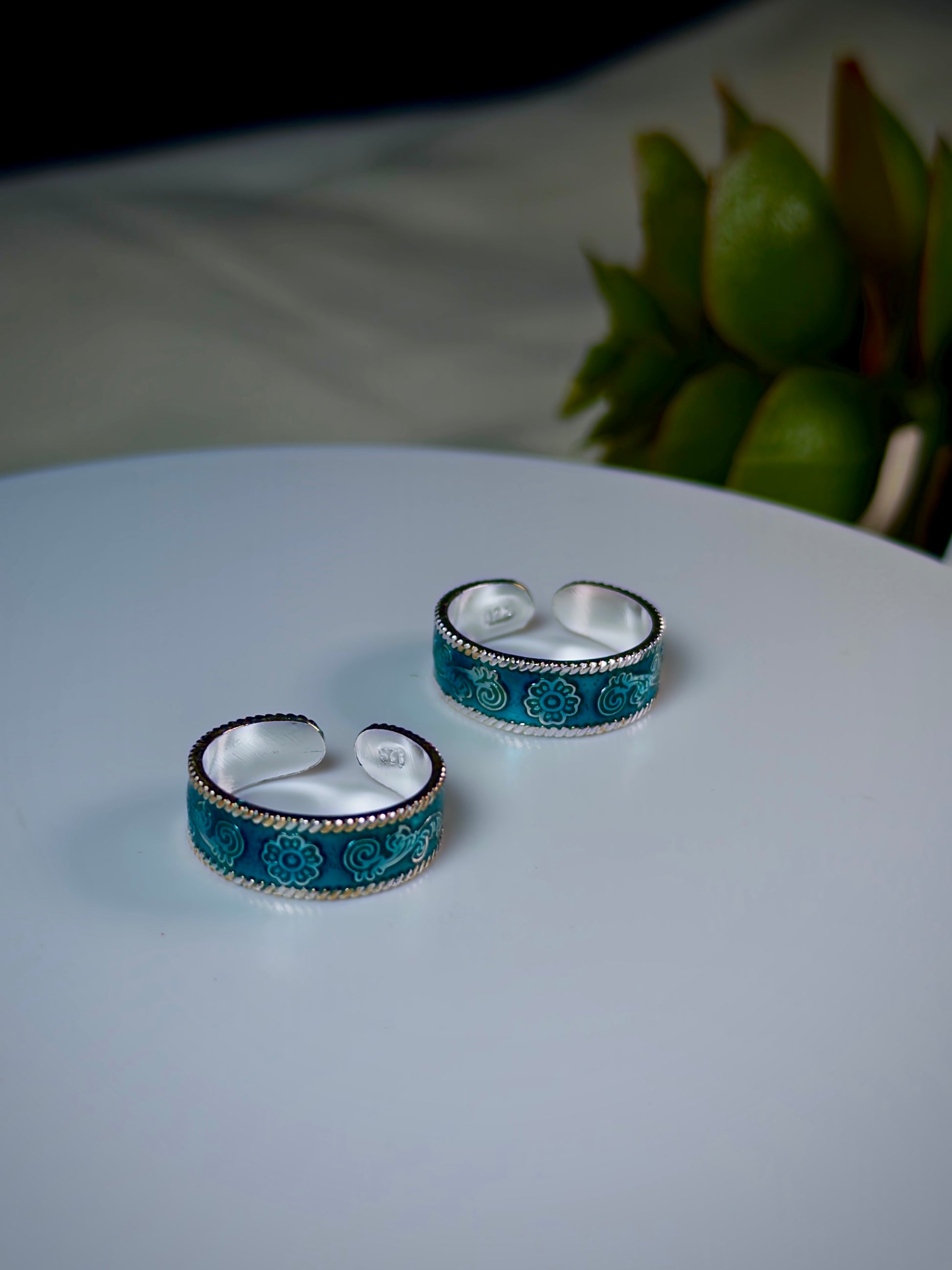Pair of blue and silver floral-patterned toe rings on a white surface with a blurred green plant in the background.