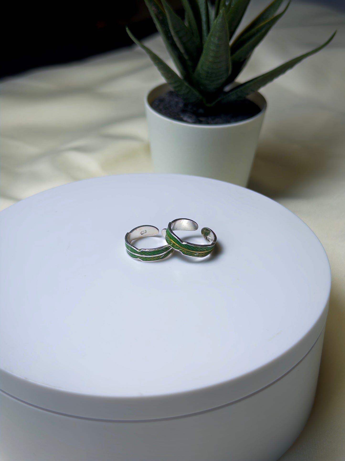 Pair of silver toe rings with green accents on a white surface, with a potted plant in the background.