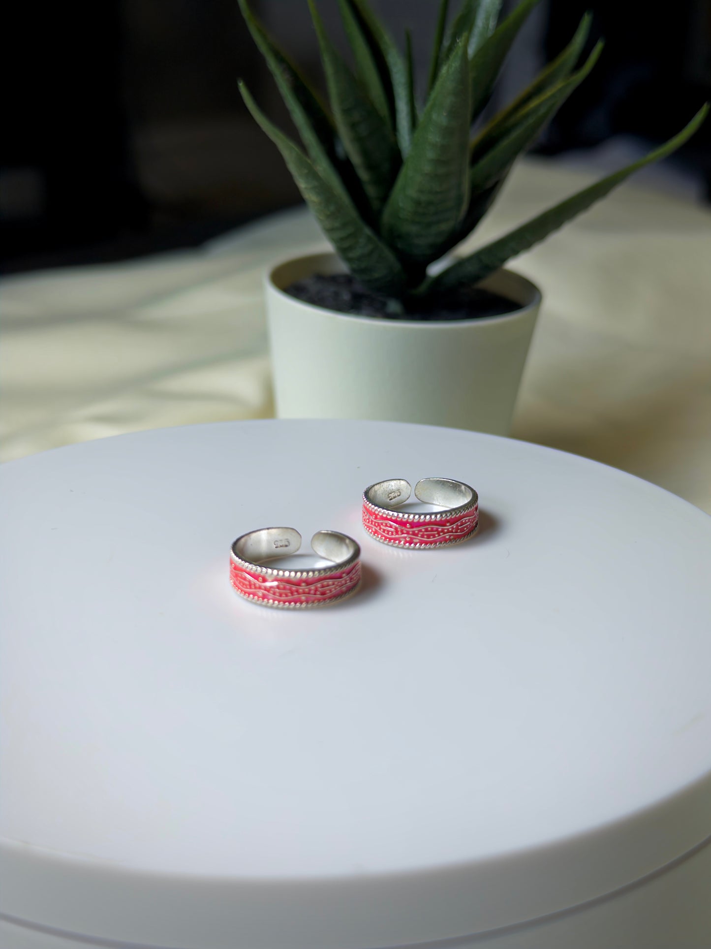 Pair of silver and pink toe rings on a white surface with a plant in the background