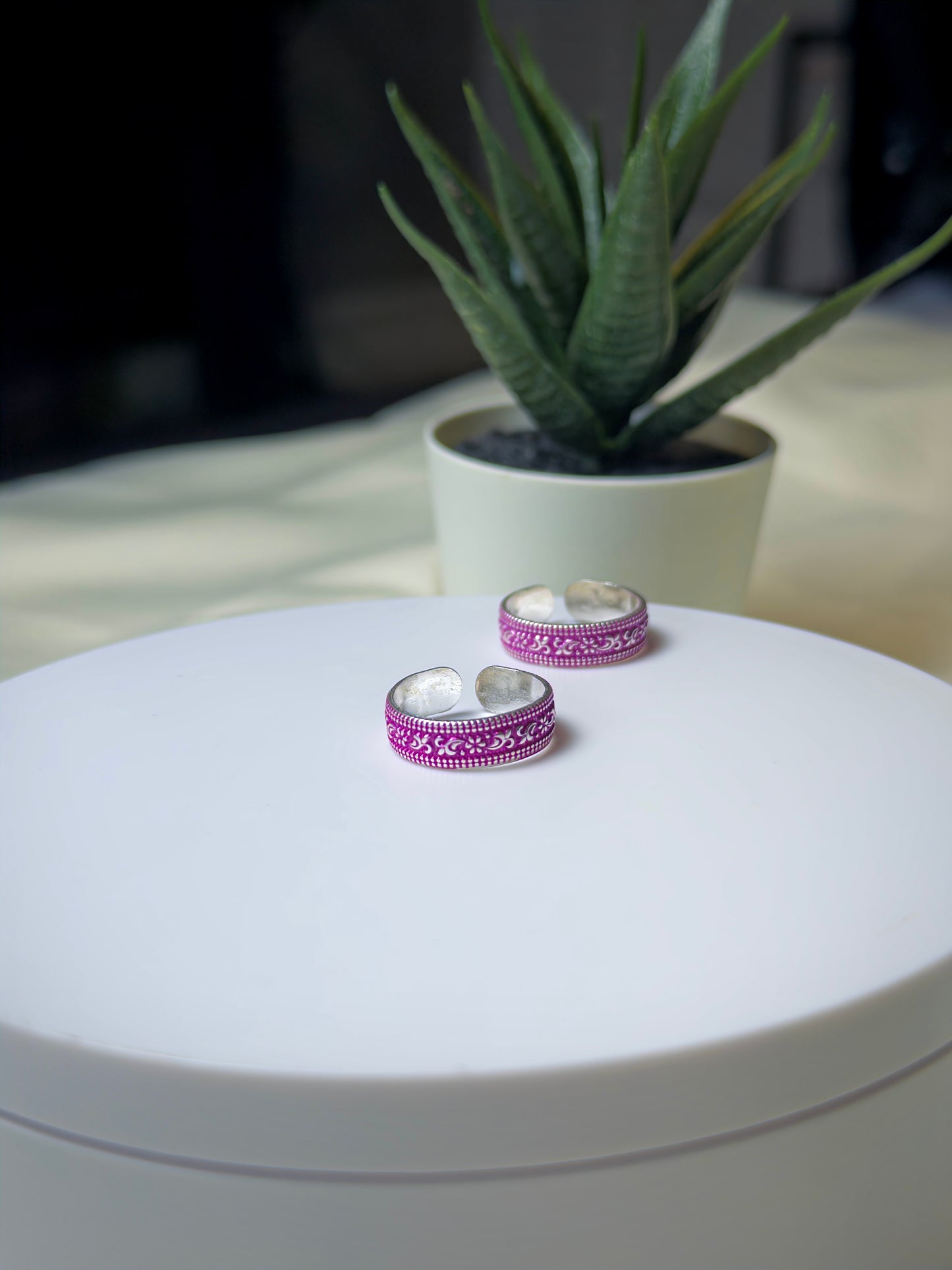 Two silver toe rings with pink patterns on a white surface, with a plant in the background.