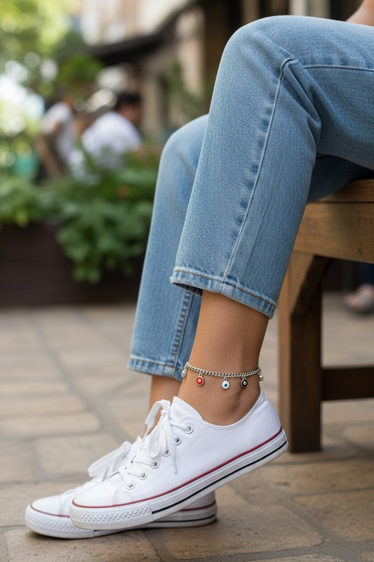 Person wearing white sneakers with evil eye anklet and blue jeans on a sidewalk.