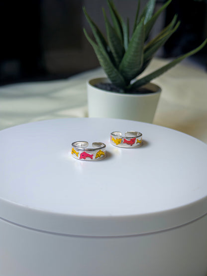 Two silver toe rings with colorful designs on a white surface, with a plant in the background.