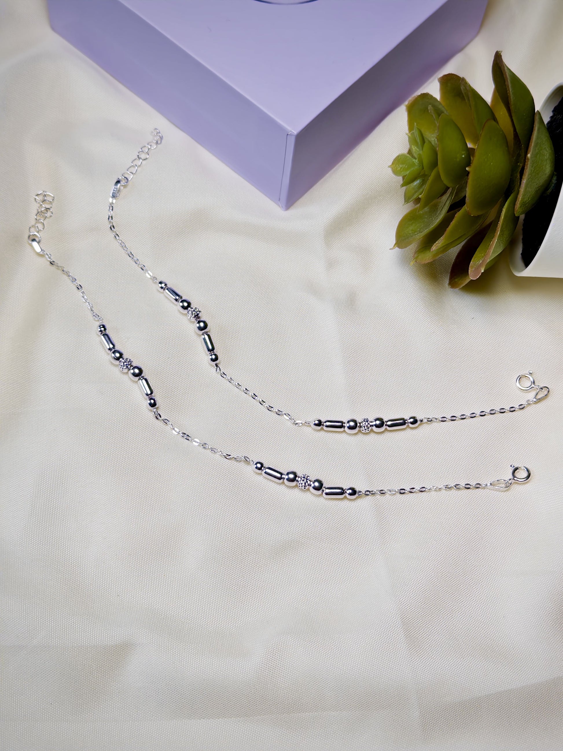 Silver beaded anklets on a light surface with a purple box and green plant in the background