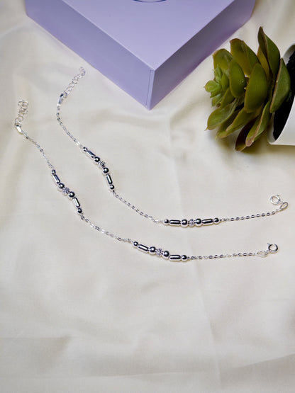 Silver beaded anklets on a light surface with a purple box and green plant in the background