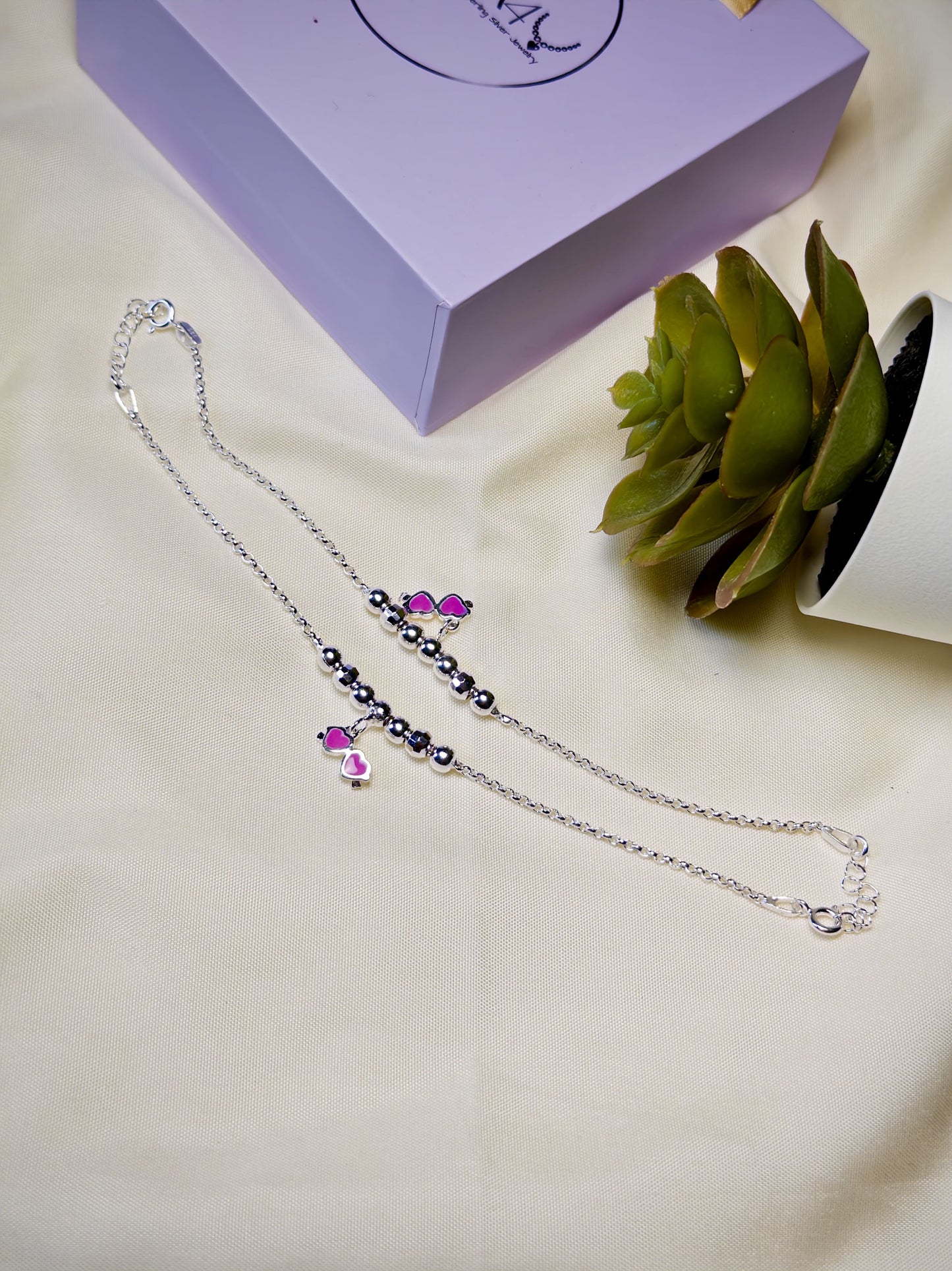Kids silver anklet with pink glasses charm on a light surface with a plant in the background