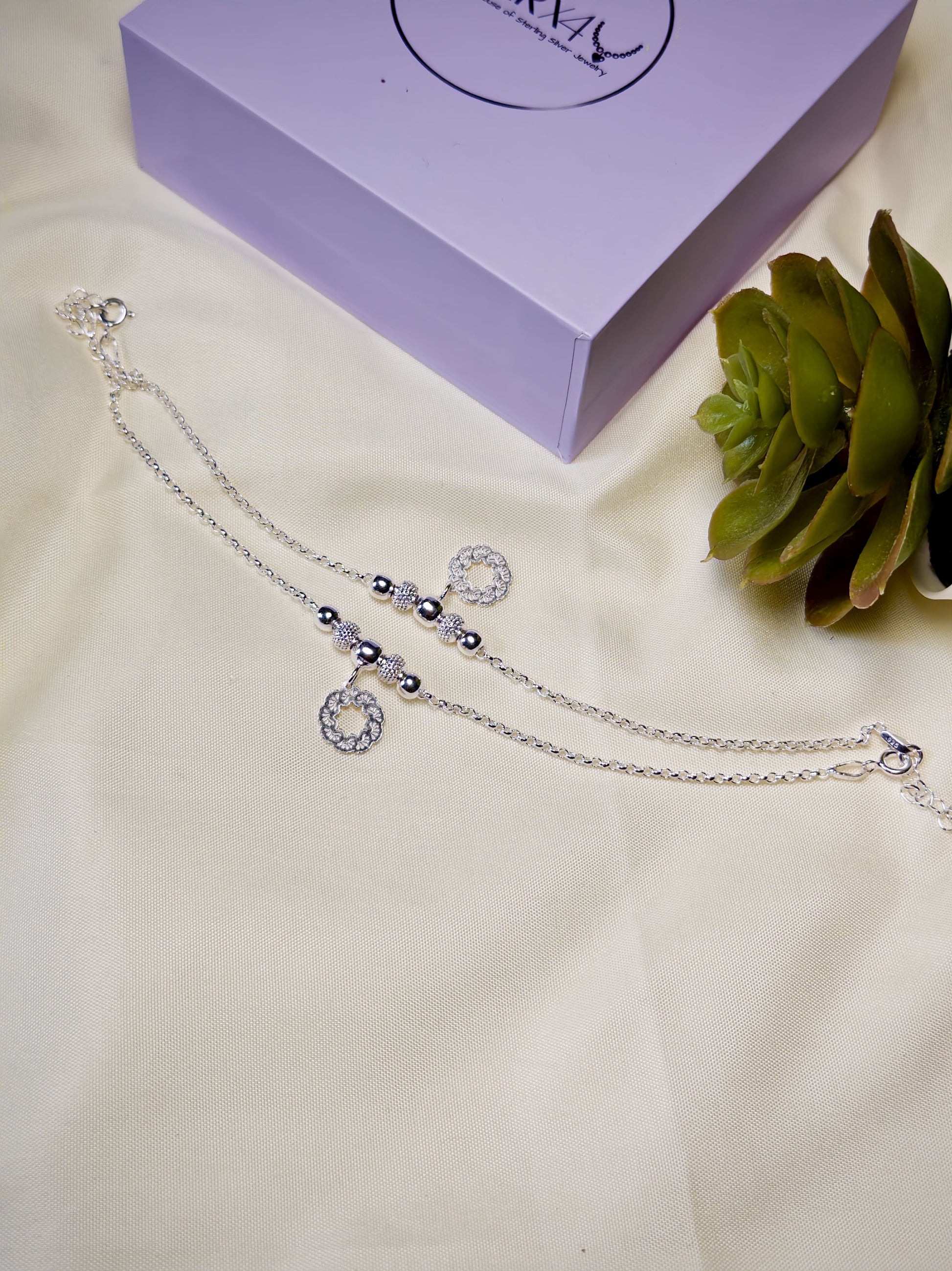 Kids silver anklets on a white surface with a purple box and green plant in the background.