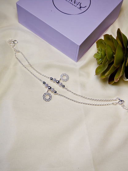 Kids silver anklets on a white surface with a purple box and green plant in the background.