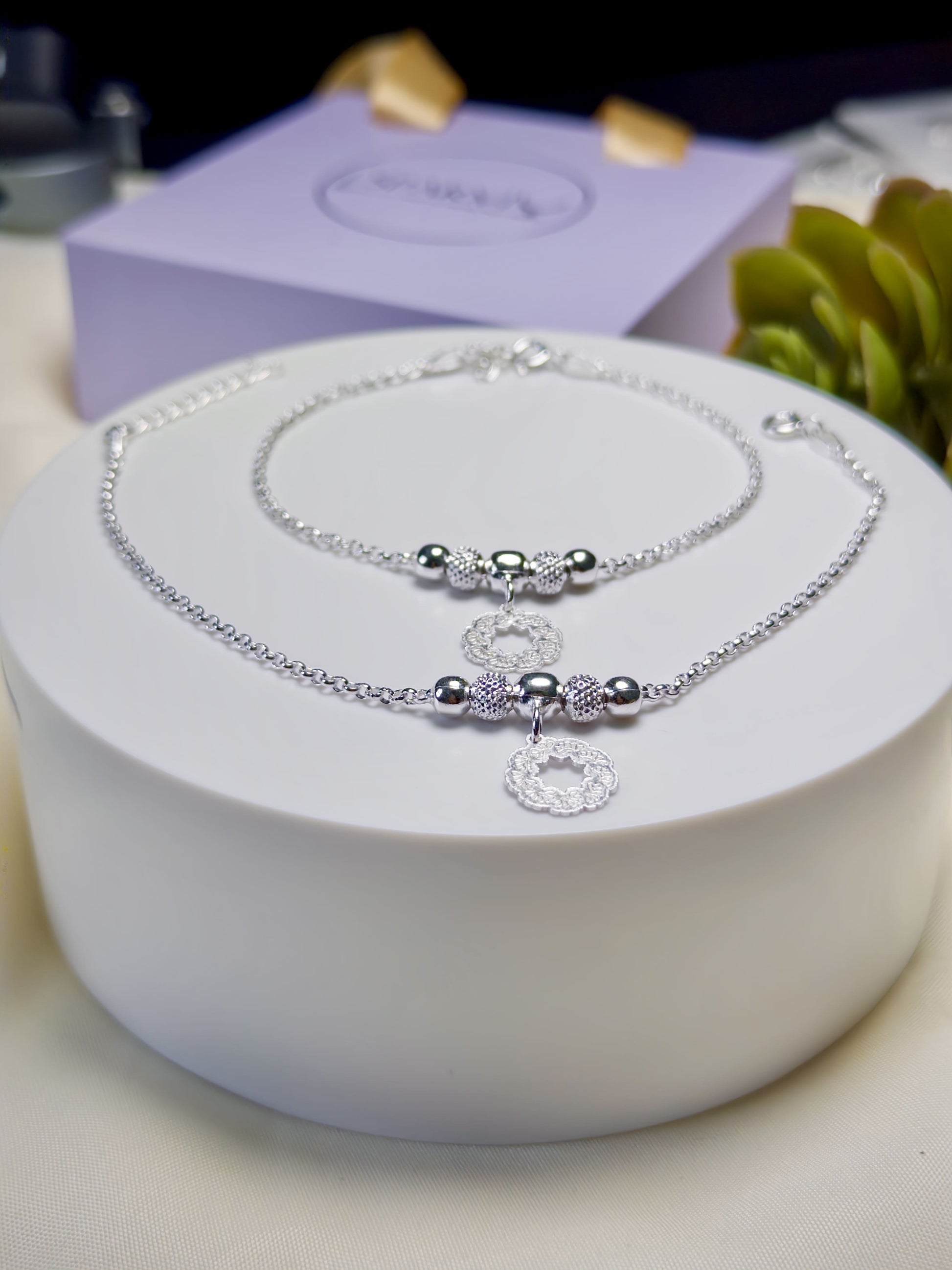 Kids silver anklets with beads on a white surface with a purple box in the background