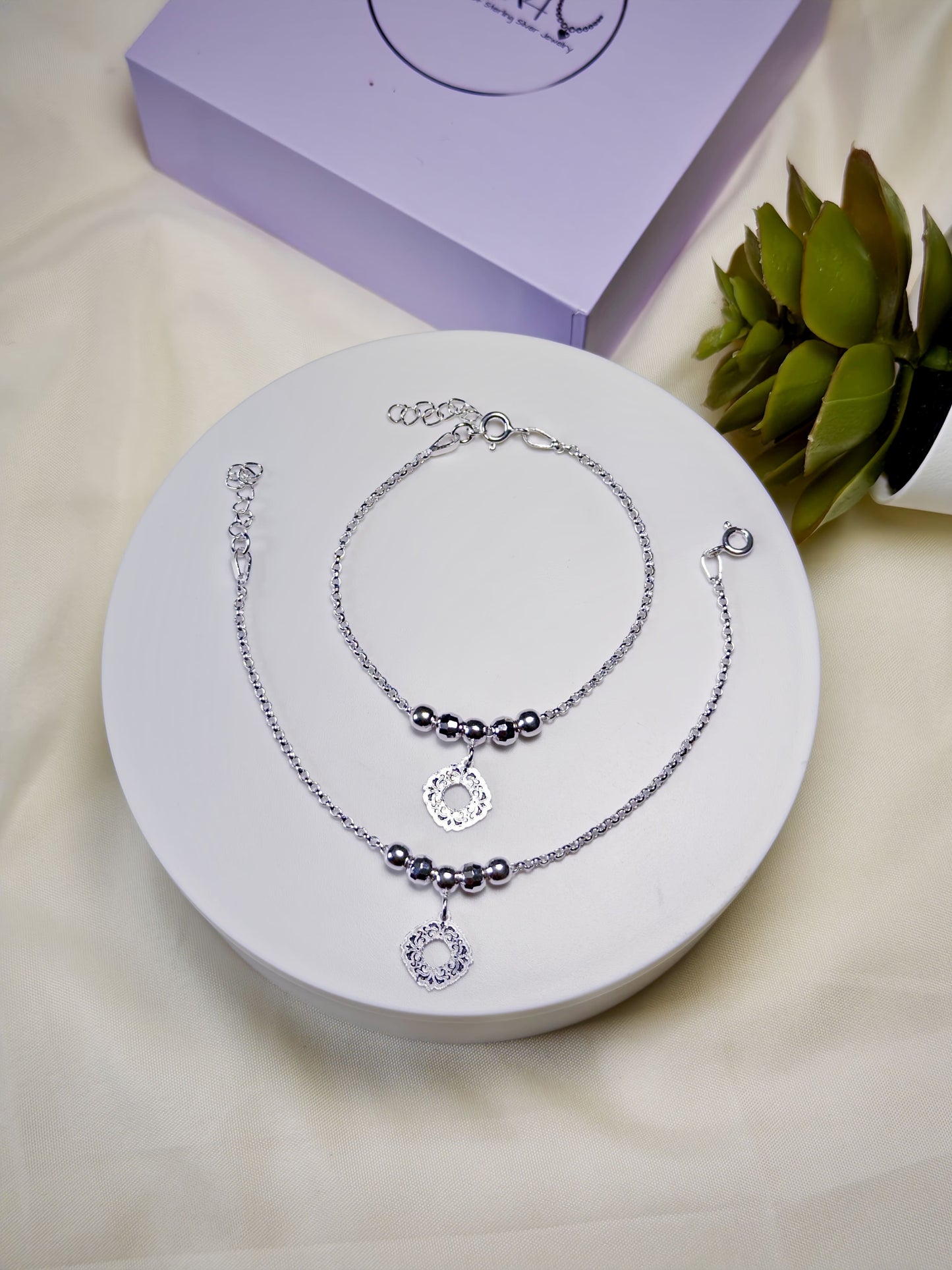 Two silver anklets with charm on a white surface, with a purple box and green plant in the background.