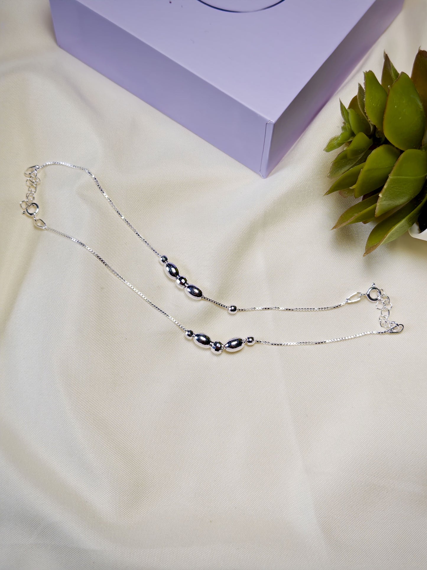 Kids silver anklets with silver beads on a white surface with a purple box and green plant in the background.