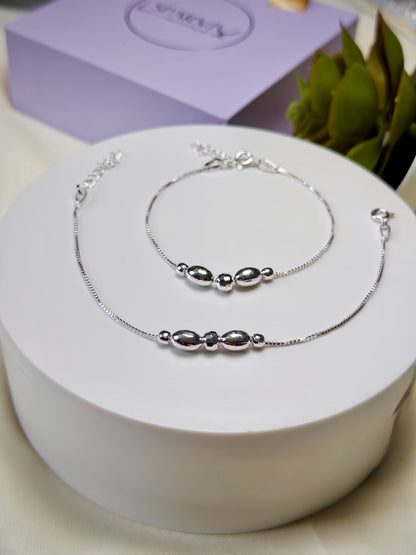 Silver anklets with silver beads on a white box with a purple box and green leaves in the background