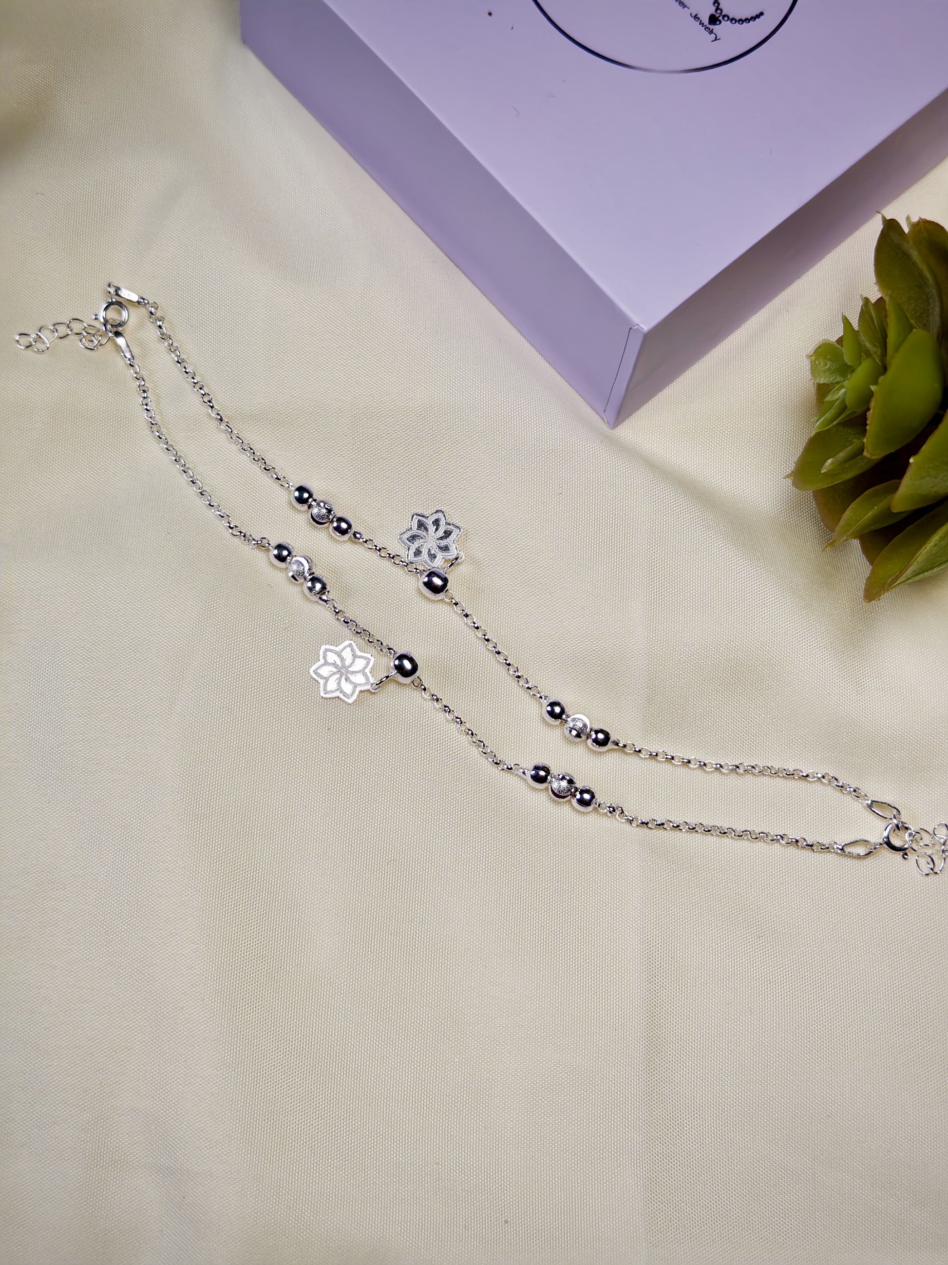 Kids silver anklets with snowflake charms on a beige surface with a purple box and green plant in the background.