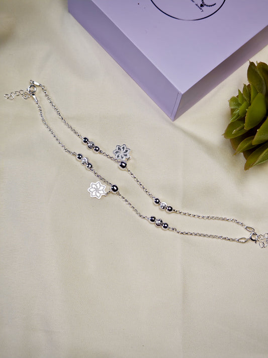 Kids silver anklets with snowflake charms on a beige surface with a purple box and green plant in the background.