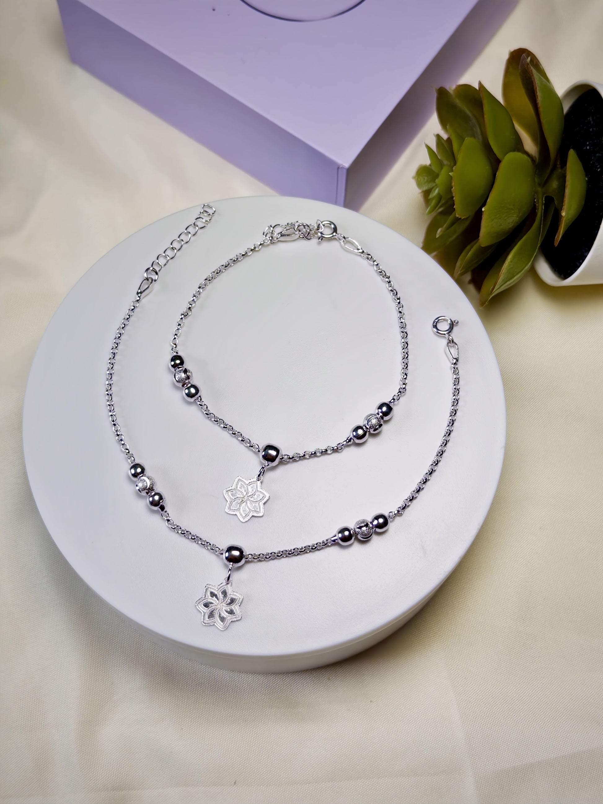 Kids silver anklets with floral pendants on a white stand with a purple box and plant in the background.