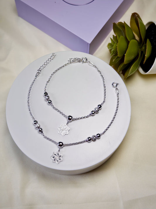 Kids silver anklets with floral pendants on a white stand with a purple box and plant in the background.