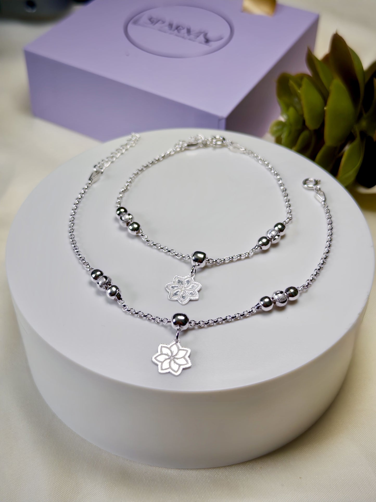 Kids silver anklets with floral charm on a white stand, with a purple box in the background.