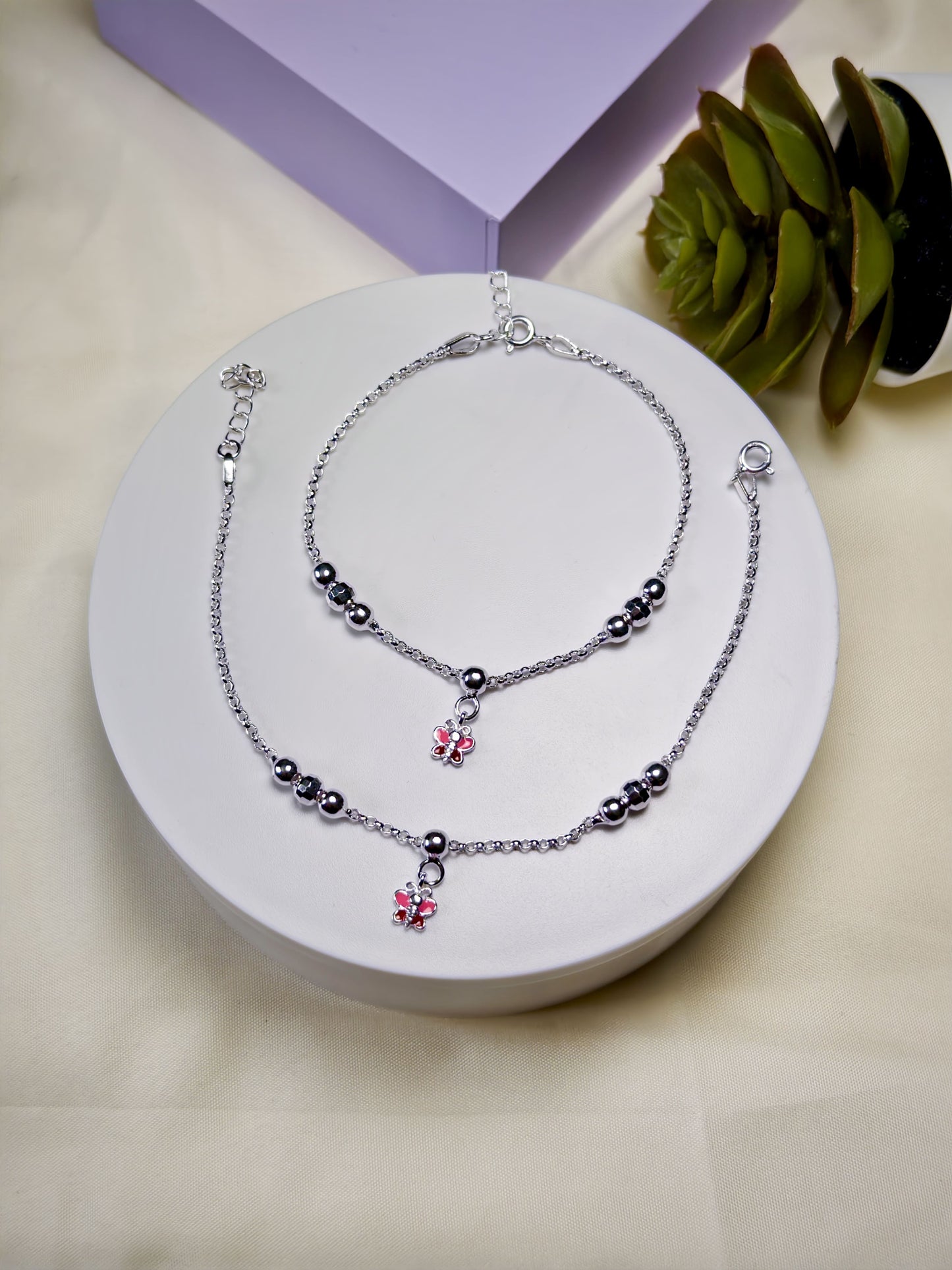 Pair of silver anklets with butterfly charm on a white surface with a purple box and green leaves in the background.