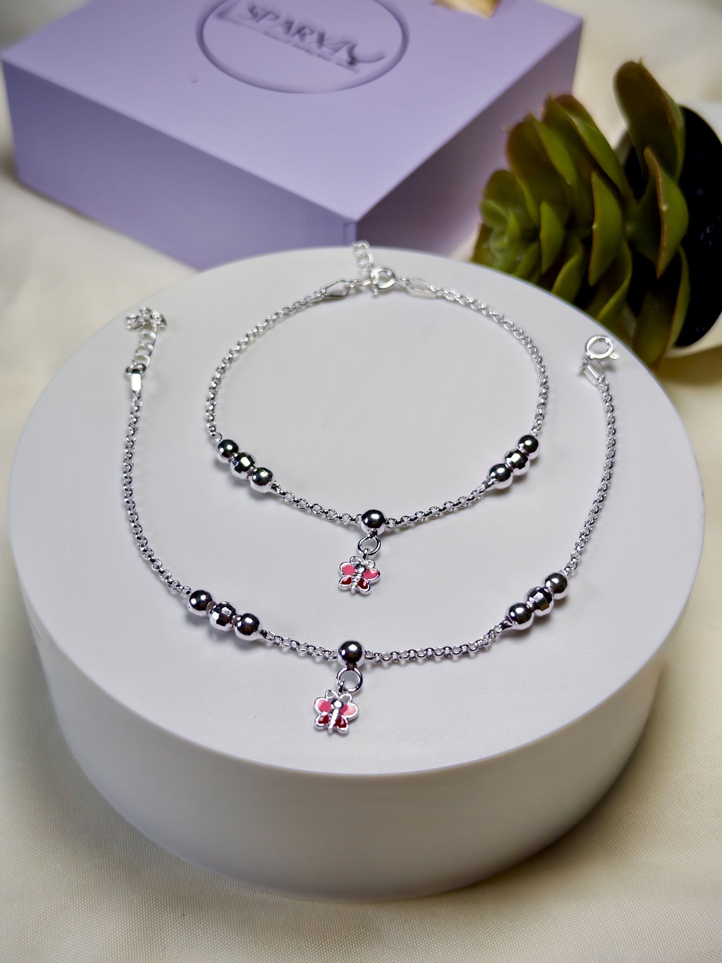 Kids silver anklets with pink butterfly charms on a white stand, with a purple box and plant in the background.