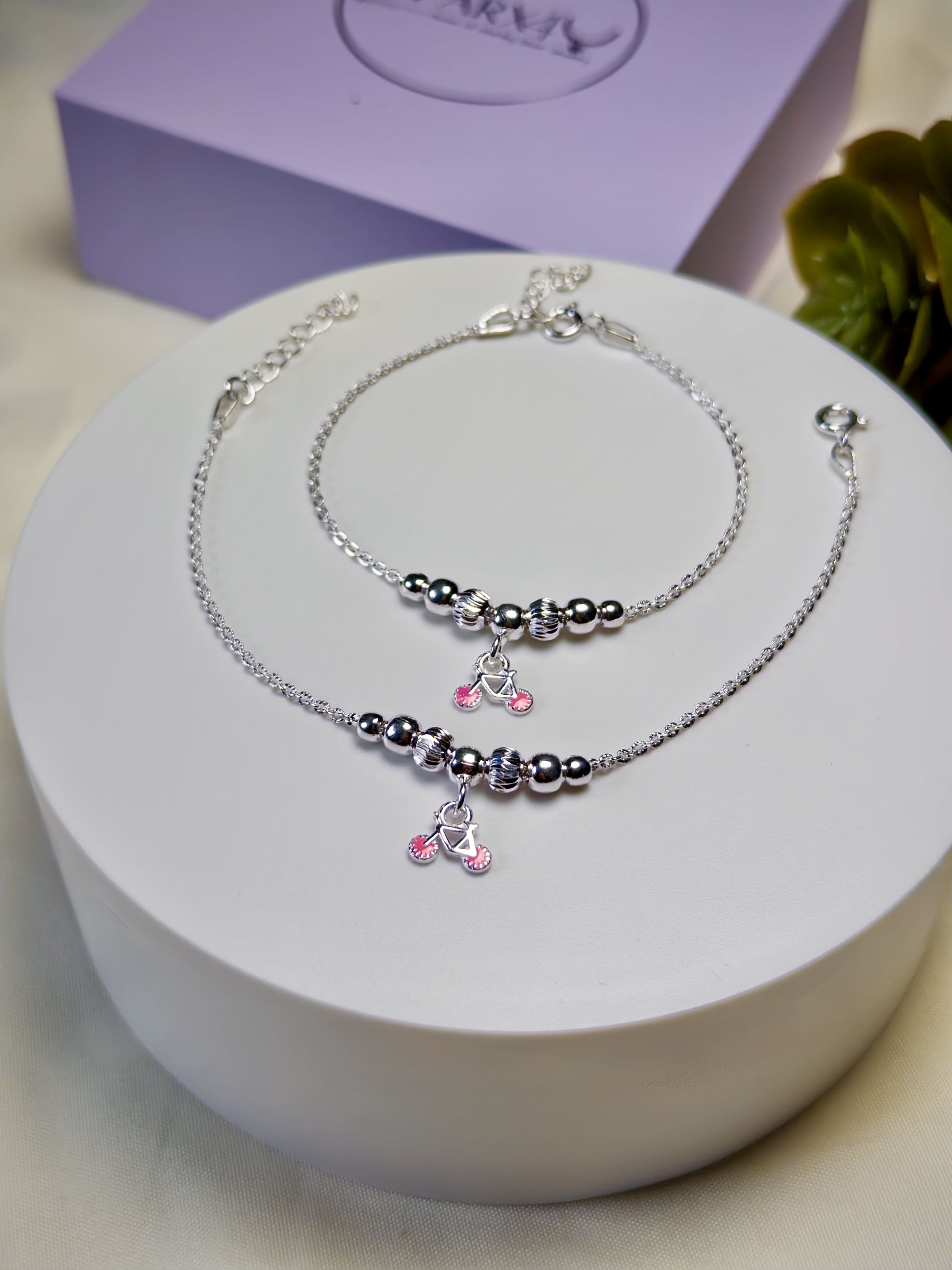 Kids silver anklets with pink cycle charms on a white surface with a purple box in the background