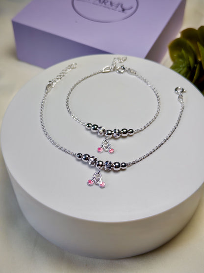 Kids silver anklets with pink cycle charms on a white surface with a purple box in the background
