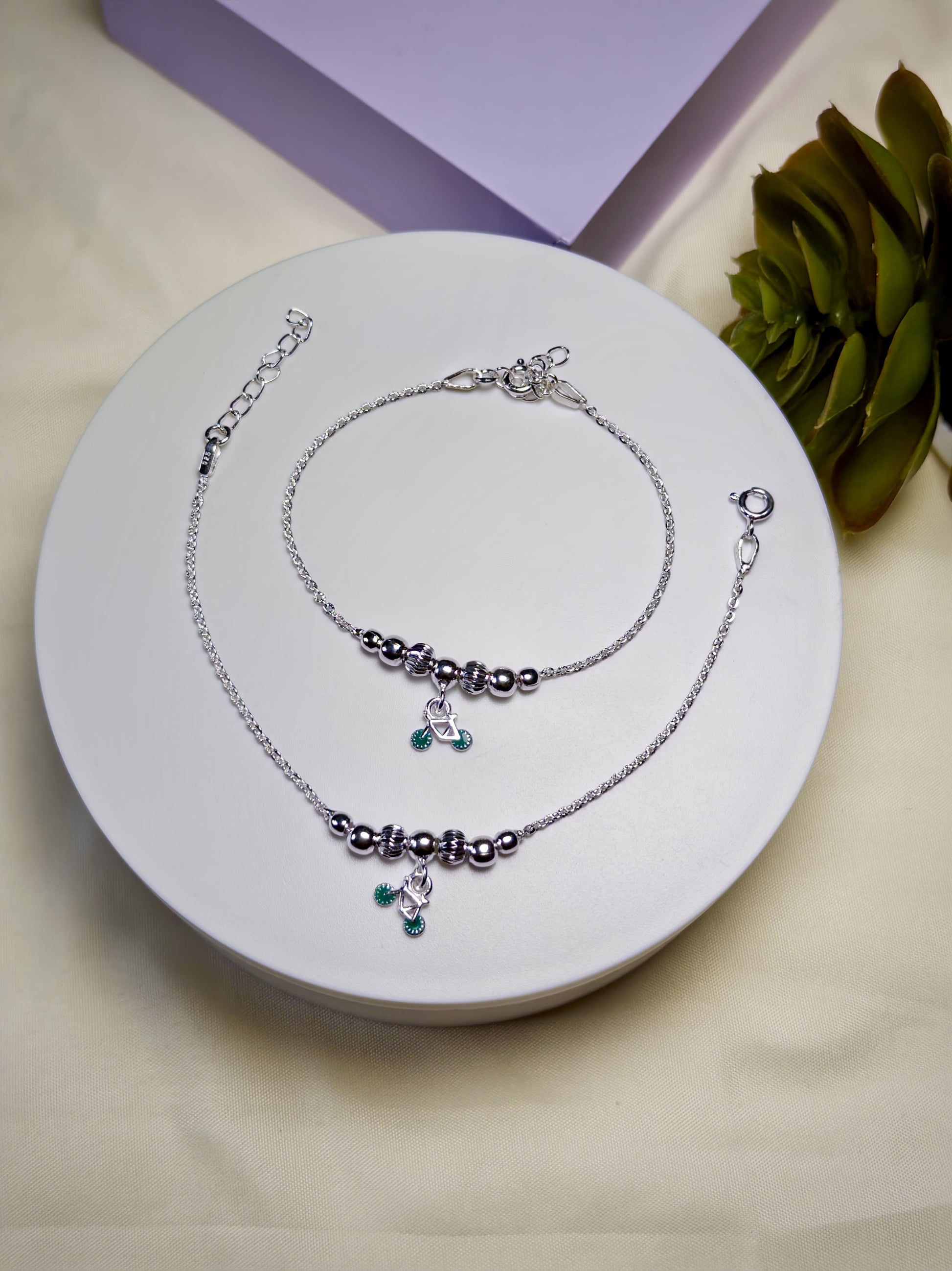 Kids silver anklets with green charms on a white plate with a purple box and green plant in the background