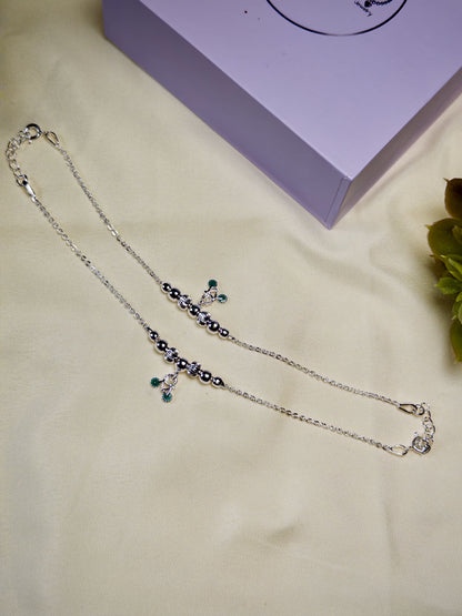 Kids silver anklets with green charm on a beige surface with a purple box in the background